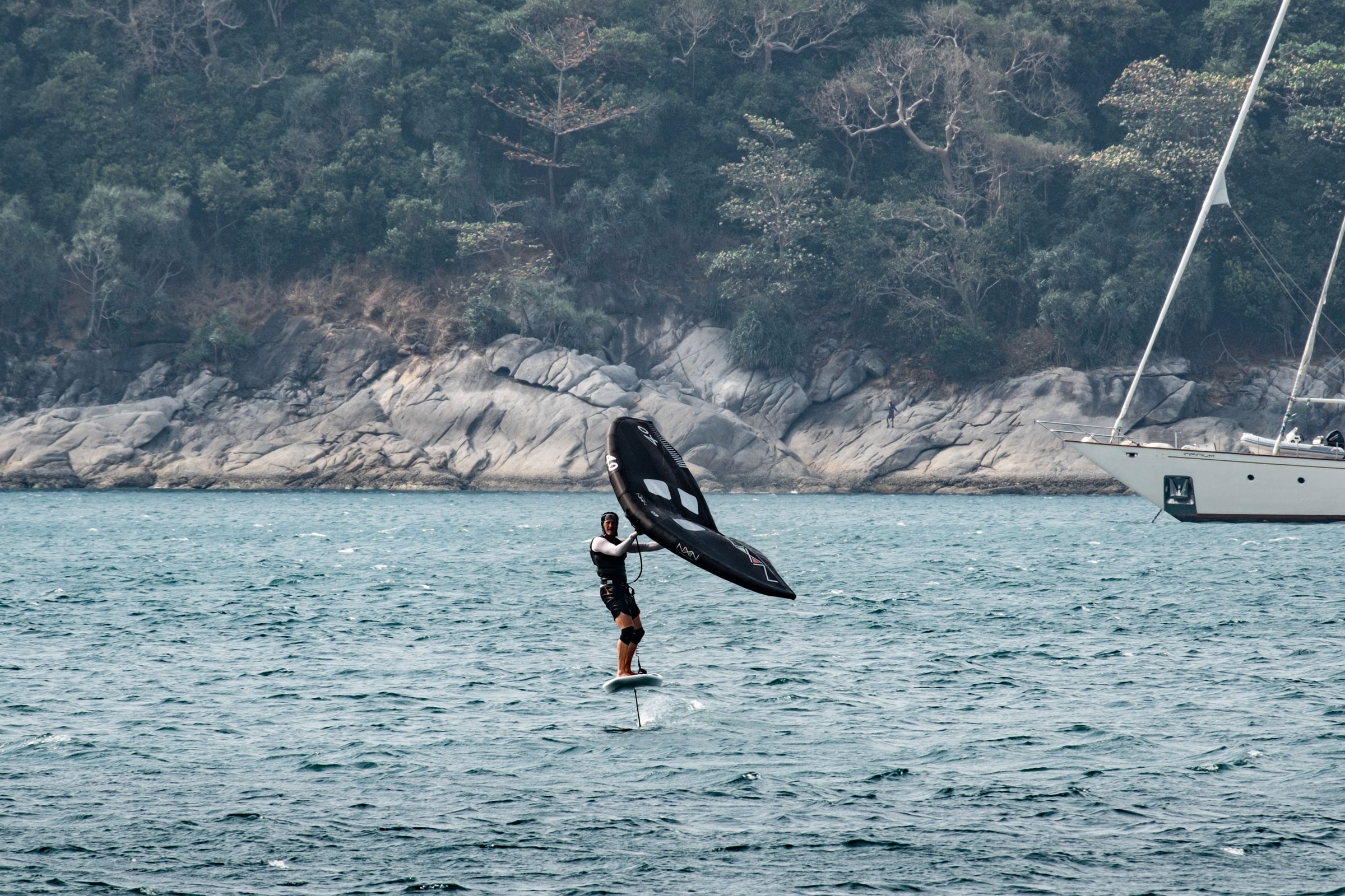 david smart riding towards the camera on a nexen foil board and wing on choppy waters. Rock cliff and trees in the background.