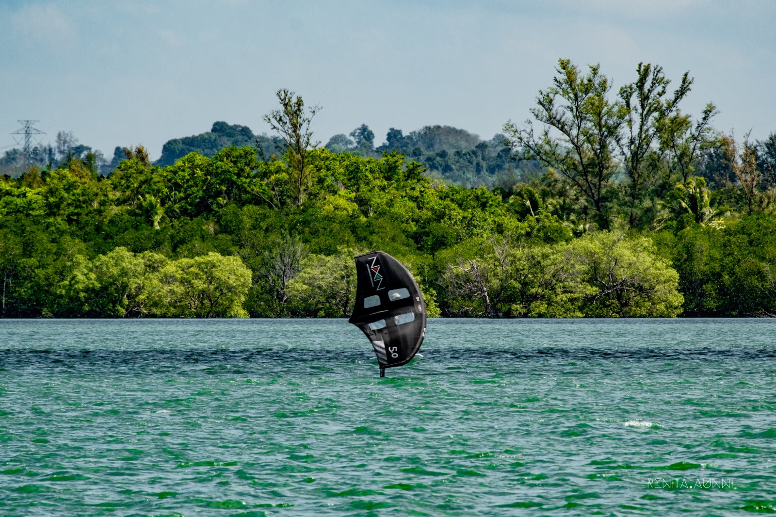 wide shot of a rider on a foil board and wing in the middle of the water with lush green trees in the background