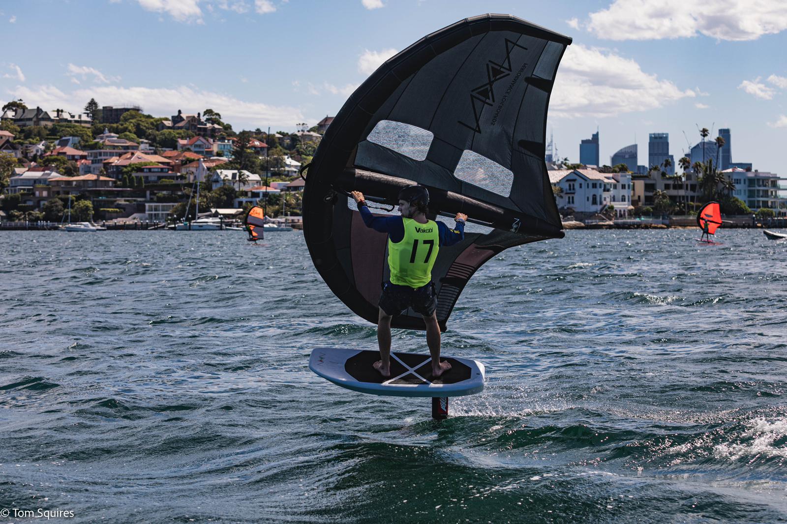 lincoln dews riding a foil board and wing in the water on a sunny day, with other riders in the background. Beachfront homes are in the background.