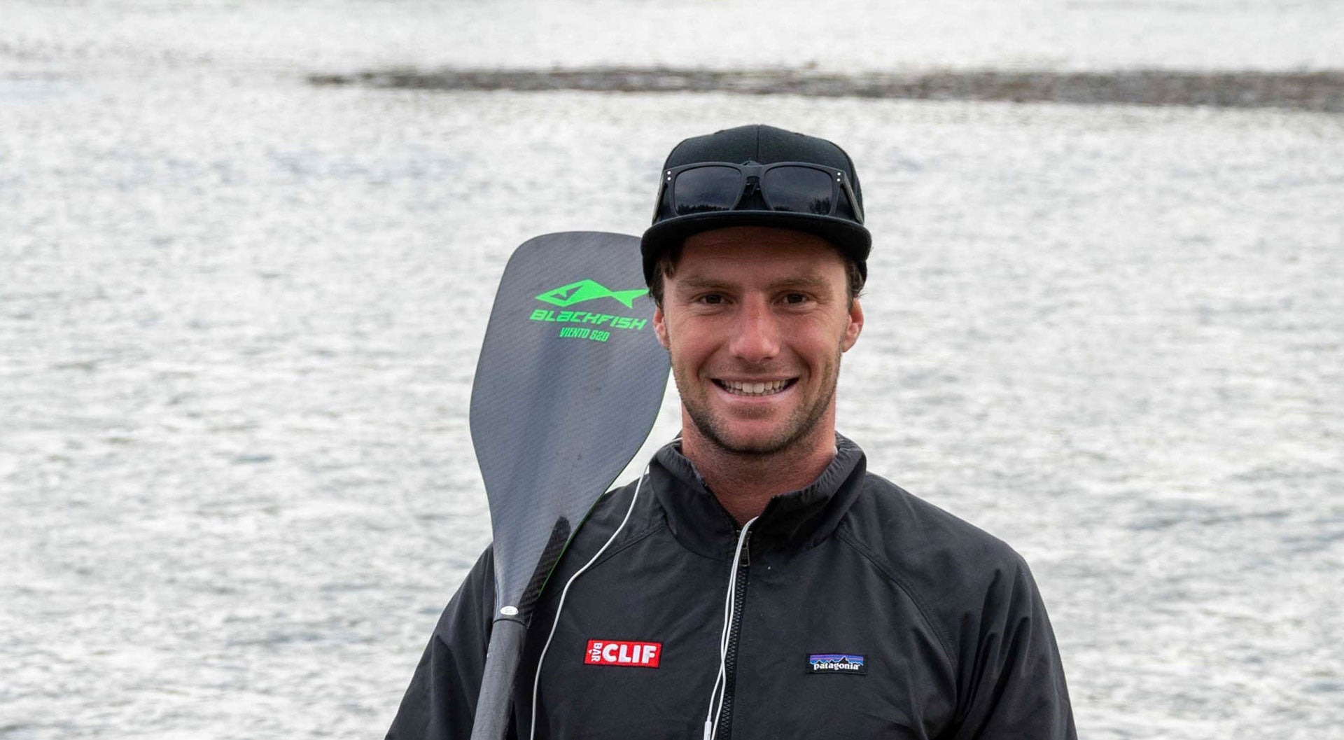 lincoln dews smiling into the camera, holding a paddle over right shoulder, with water in the background