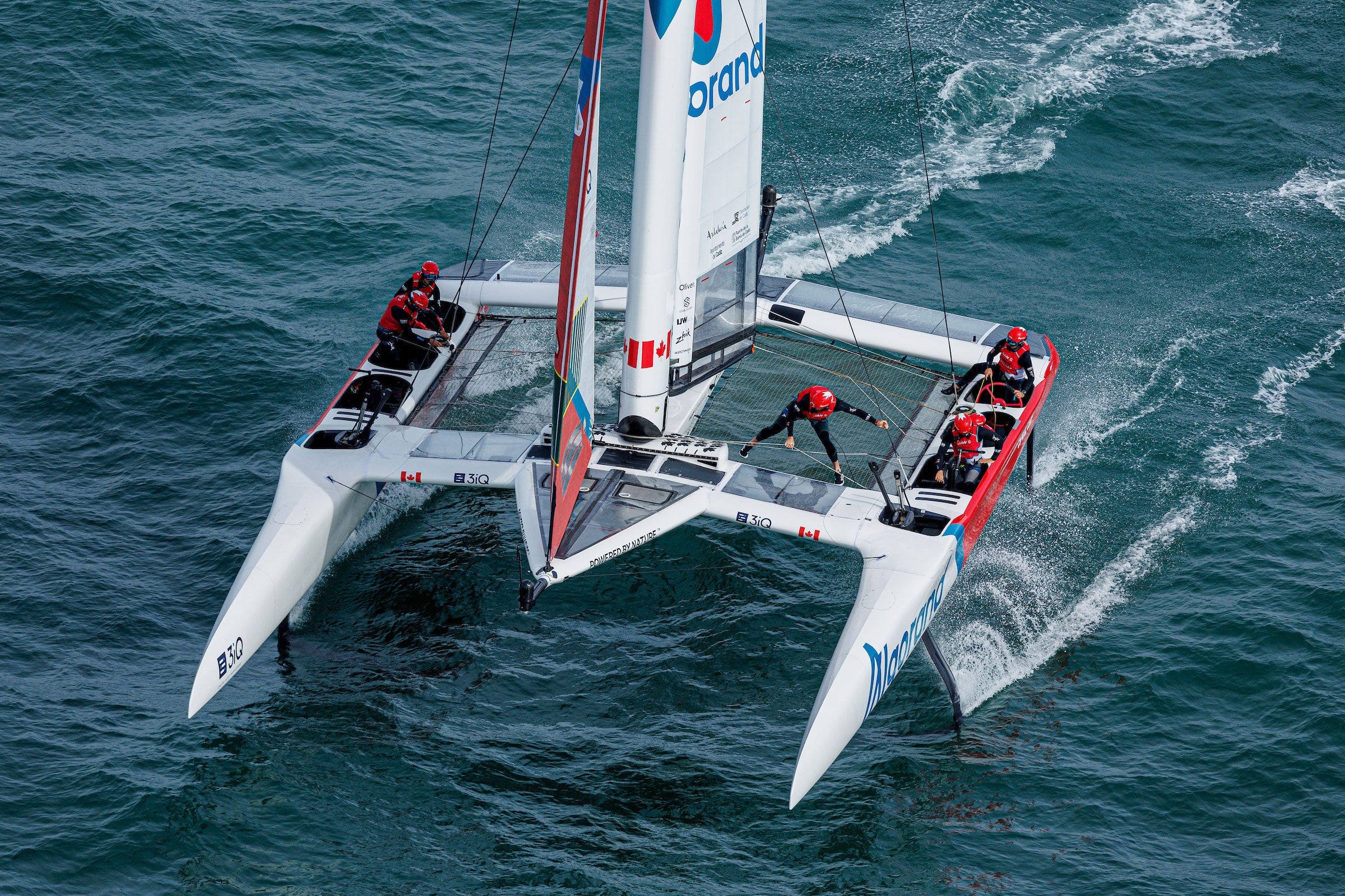 top down view of the SailGP Canada boat sailing through waters, showing the crew in their seats sailing the boat, with one crew member standing, handling the sail.