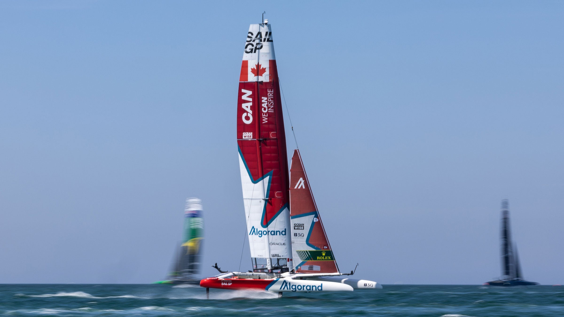 side view of the SailGP Canada boat racing through the waters, with blurred boats in the background, showing the speed of the boat.