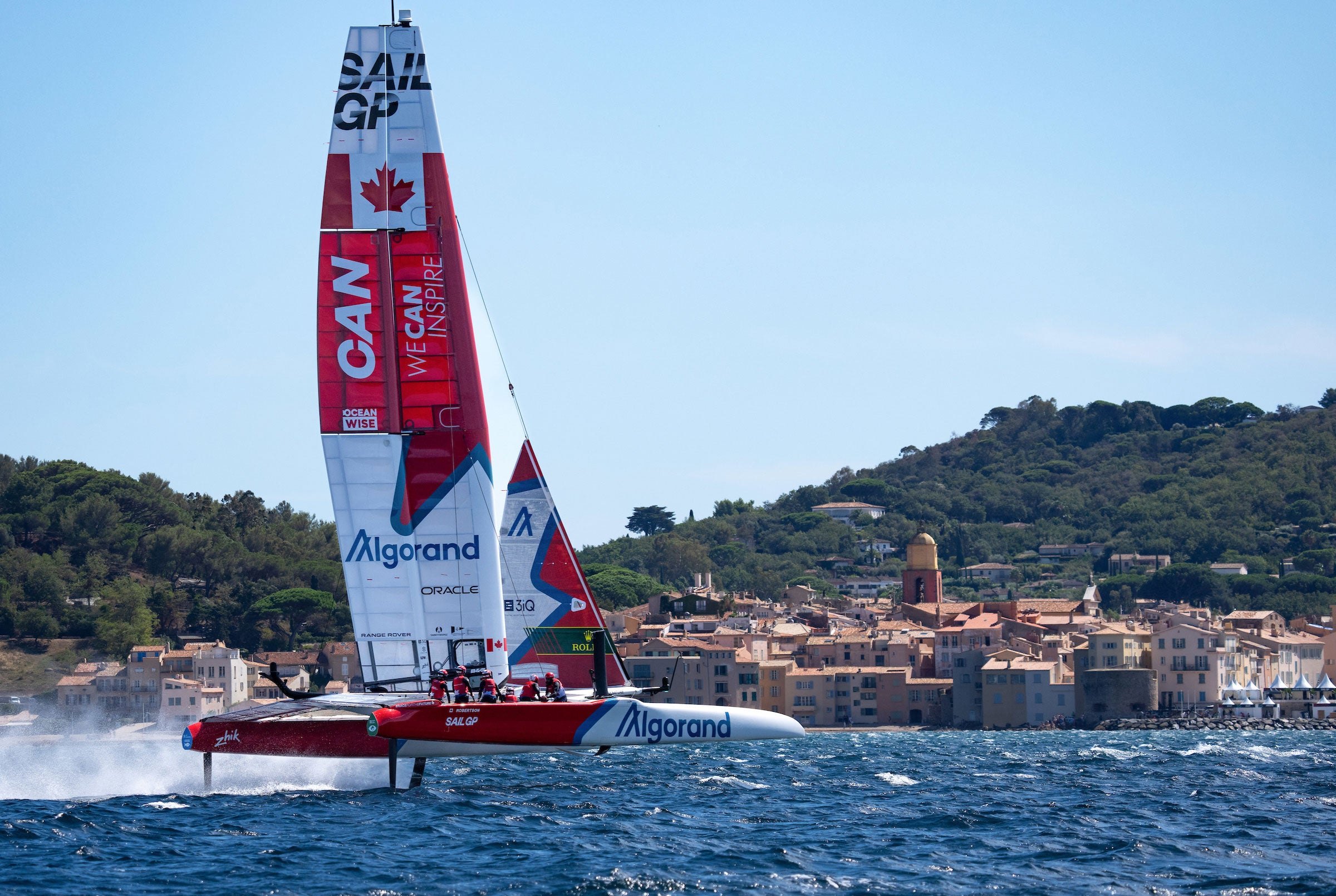 SailGP Canada boat sailing rapidly through choppy waters, with green hills and beach front home in the background